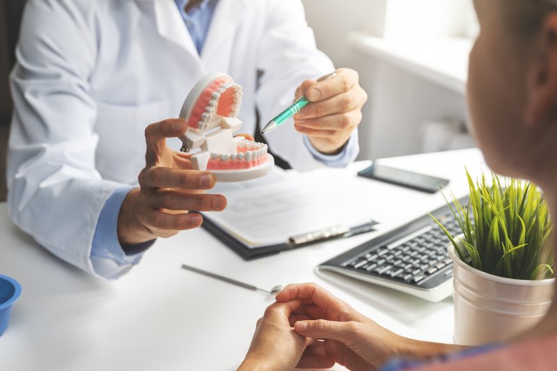 Dentist pointing pen at model of teeth that's opened wide to woman sitting across desk from them